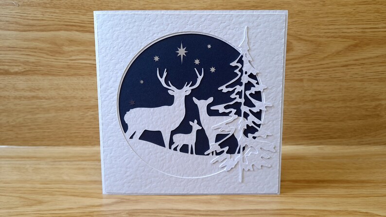 May include: A white card with a blue circle cut out in the center. The circle features a silhouette of three deer in a snowy forest with stars in the night sky. The card is decorated with a white silhouette of a pine tree.