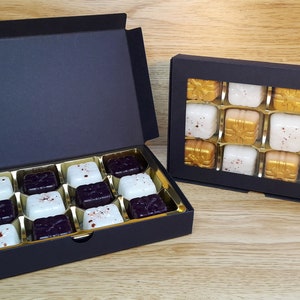 May include: Two black gift boxes with lids open, revealing rows of square chocolates. The chocolates are arranged in a grid pattern and are white, gold, and dark purple. The boxes are on a wooden surface.