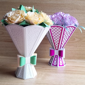 Op de afbeelding: Twee decoratieve papieren bloemboeketten. Eén boeket heeft crèmekleurige papieren rozen in een witte vaas met een groene strik. Het andere boeket heeft paarse papieren bloemen in een magenta en witte vaas met een magenta strik.