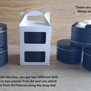 May include: Two black metal tins with gold trim, stacked on top of each other. The tins are 8 ounces and measure 80mm wide and 53mm tall. A white cardboard box with a handle and two cut-out windows is also shown. The text "These are 8oz tins which are 80mm wide and 53mm tall" is visible in the image.