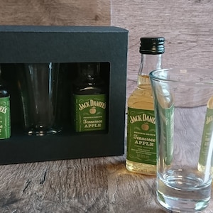 May include: A black gift box with two small bottles of Jack Daniel's Tennessee Apple liqueur and two shot glasses. The bottles are green and have a gold label with the Jack Daniel's logo and the words "Tennessee Apple".
