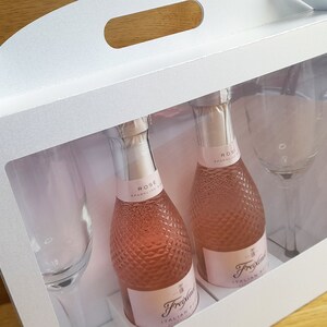 May include: A white gift box with a handle and a clear window. The box contains two small bottles of pink sparkling wine and two champagne flutes. The bottles are labeled "Rose Sparkling Wine" and "Fragolino Italian Sparkling Wine".