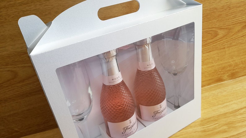 May include: A white gift box with a handle and a clear window. The box contains two small bottles of pink sparkling wine and two champagne flutes. The bottles are labeled "Rose Sparkling Wine" and "Fragolino Italian Sparkling Wine".