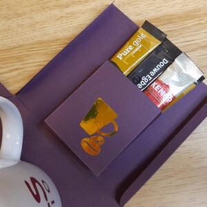 May include: A purple gift box with a white mug and gold accents. The box has a pocket on the side with three coffee packets: Pure Gold, Double Edge, and Kenco.