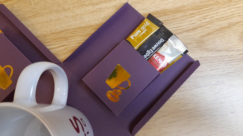 May include: A purple gift box with a white mug and gold accents. The box has a pocket on the side with three coffee packets: Pure Gold, Double Edge, and Kenco.