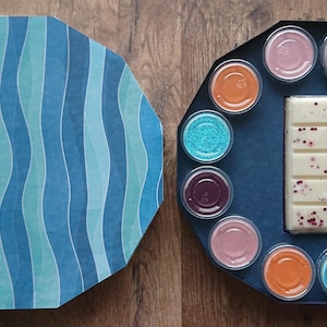 May include: A round, blue and green patterned box with a lid open to reveal a white chocolate bar with red sprinkles and a circle of small, colorful wax melts.