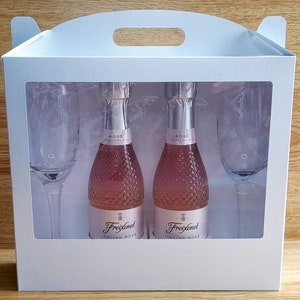 Può includere: Una scatola regalo bianca con una finestra trasparente che mostra due piccole bottiglie di vino spumante rosato italiano e due flute da champagne.