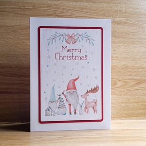 Pode incluir: Um cartão de Natal com uma borda vermelha apresenta as palavras "Merry Christmas" acima de ilustrações de gnomos e uma rena. Os gnomos usam chapéus azuis, cinzentos e vermelhos. A rena é castanha clara. O cartão está em uma superfície de madeira.