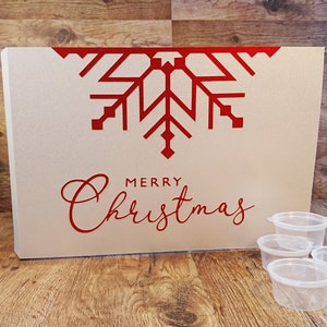 Könnte beinhalten: Eine rechteckige, cremefarbene Box mit einem roten Schneeflocken-Design und dem Schriftzug "MERRY Christmas" in Rot. Mehrere kleine, transparente Kunststoffbehälter befinden sich vor der Box. Die Box steht auf einer Holzoberfläche.