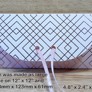 May include: A white gift box with a geometric pattern in rose gold. The box has a rounded flap and is tied with a pink ribbon. The box measures 4.8 inches by 2.4 inches by 2.4 inches.