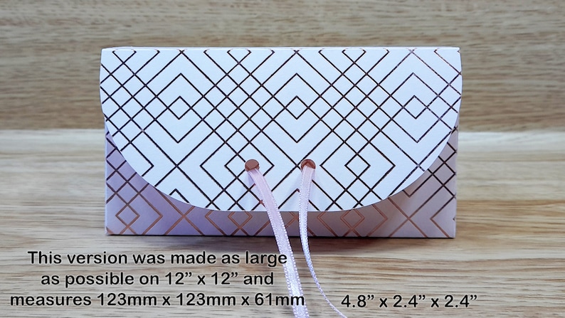 May include: A white gift box with a geometric pattern in rose gold. The box has a rounded flap and is tied with a pink ribbon. The box measures 4.8 inches by 2.4 inches by 2.4 inches.