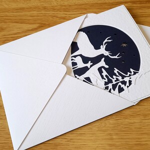 May include: A white envelope with a blue and white greeting card inside. The card features a laser-cut design of deer and trees against a dark blue background.