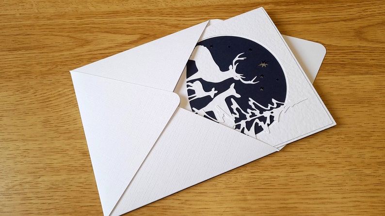 May include: A white envelope with a blue and white greeting card inside. The card features a laser-cut design of deer and trees against a dark blue background.