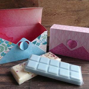 May include: Two decorative boxes with a heart-shaped opening on the front. The boxes are made of paper and have a floral pattern. The boxes are open and contain two bars of wax melt. One bar is white with red flecks and the other is light blue.