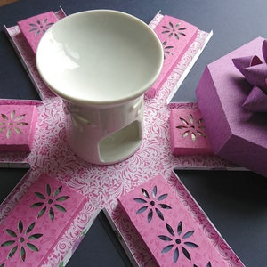 May include: A white ceramic wax warmer with a decorative floral design on the base. The warmer is surrounded by pink paper boxes with cut-out floral designs. A purple gift box with a bow is also included.