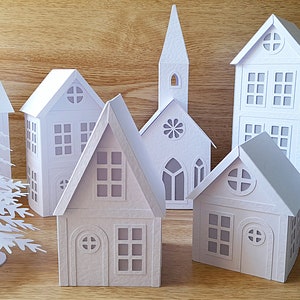 Puede incluir: Un conjunto de casas y árboles de papel blanco, que incluye una iglesia, una casa alta y dos casas más pequeñas. Las casas tienen ventanas y puertas recortadas, y los árboles tienen un diseño delicado y de encaje.