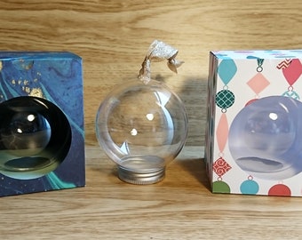 Op de afbeelding: Drie heldere glazen kerstballen met zilveren toppen, elk in een andere doos. De dozen zijn versierd met verschillende patronen, waaronder een blauw en gouden marmerpatroon, een witte achtergrond met kleurrijke geometrische vormen en een witte achtergrond met kleurrijke kerstballen.