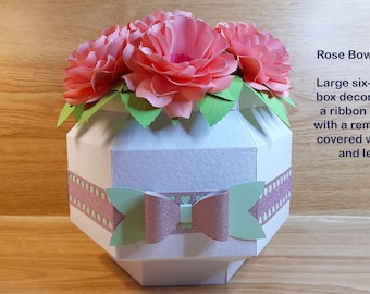 May include: A white six-sided gift box decorated with a pink ribbon and bow, covered with pink paper roses and green leaves. The box has a removable lid. "Rose Bowl Gift Box" is written in the image.