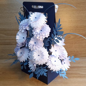 May include: A blue paper bag filled with a bouquet of white paper flowers and blue paper leaves.