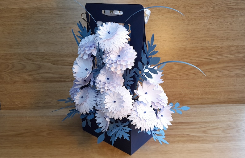May include: A blue paper bag filled with a bouquet of white paper flowers and blue paper leaves.