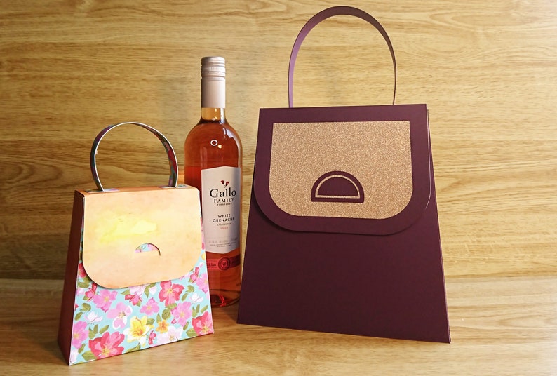 Pode incluir: Duas sacolas de presente com al&ccedil;as, uma com um padr&atilde;o floral e a outra com um design marrom e dourado. Uma garrafa de vinho branco Grenache da Gallo Family Vineyards est&aacute; no fundo.