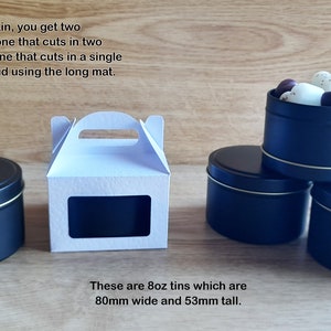 May include: A white cardboard box with a window and a handle, next to three black metal tins. The tins are 8 ounces and measure 80mm wide and 53mm tall. The text on the image reads: "For this box for one tin, you get two different SVG files, one that cuts in two pieces from A4, or one that cuts in a single piece from A3/Tabloid using the long mat. These are 8oz tins which are 80mm wide and 53mm tall."
