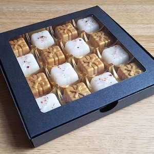 May include: A black box with a clear window showing 16 small, white and gold gift-shaped candles. The candles are arranged in a grid pattern.