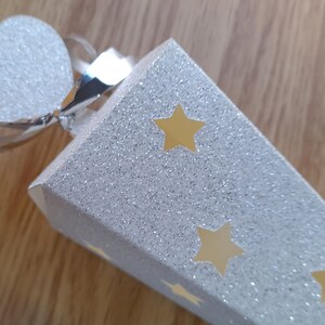 May include: A white cone-shaped party favor with a silver glitter finish and gold star cutouts. The cone has a silver ribbon and a white glitter heart attached to the top.