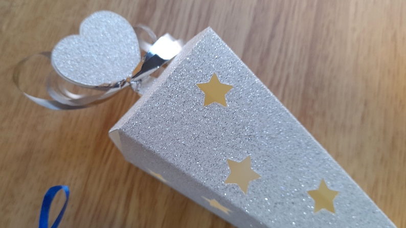 May include: A white cone-shaped party favor with a silver glitter finish and gold star cutouts. The cone has a silver ribbon and a white glitter heart attached to the top.