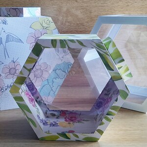 Op de afbeelding: Drie zeshoekige dozen met verschillende ontwerpen. De doos links heeft een bloemenpatroon met vogels. De middelste doos heeft een bloemenpatroon met groene bladeren. De doos rechts is wit en heeft een helder acrylvenster.