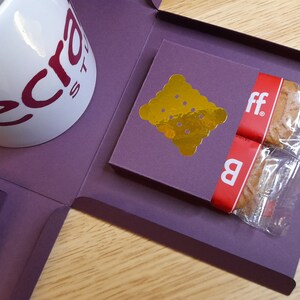 May include: A purple gift box with a white mug that says "Electra" in red letters. The box is open and contains two individually wrapped cookies with a gold foil sticker on one of the boxes.