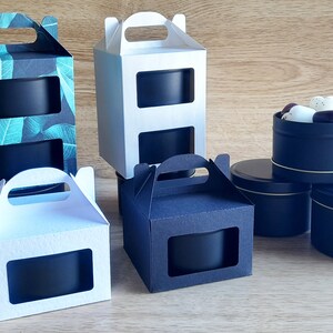 May include: Four black candle tins with clear windows, two in white boxes with handles, one in a black box with a handle, and one in a box with a green leaf pattern. The candle tins are stacked on a wooden surface.
