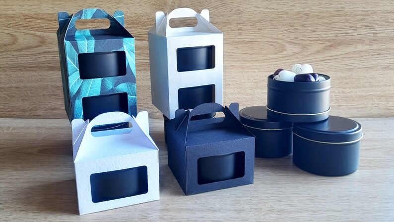 May include: Four black candle tins with clear windows, two in white boxes with handles, one in a black box with a handle, and one in a box with a green leaf pattern. The candle tins are stacked on a wooden surface.