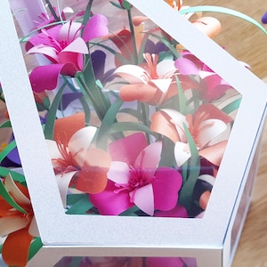 May include: A white geometric box with a clear window, filled with a bouquet of paper flowers in shades of pink, orange, and white. The flowers are arranged in a cluster, with green paper leaves.