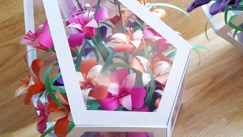 May include: A white geometric box with a clear window, filled with a bouquet of paper flowers in shades of pink, orange, and white. The flowers are arranged in a cluster, with green paper leaves.
