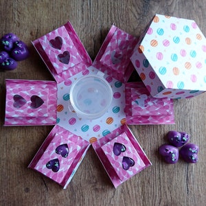 May include: A pink and white explosion box with a hexagonal lid. The box has a heart-shaped cut-out on each side and is decorated with a pattern of hearts and polka dots. The lid is decorated with a pattern of colorful circles. The box is open and there are purple heart-shaped wax melts inside.