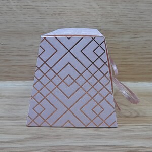 May include: A white gift box with a geometric pattern in rose gold. The box has a ribbon tied around it.