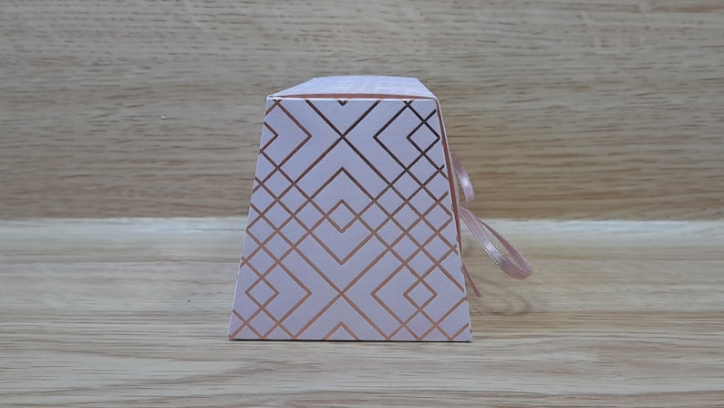 May include: A white gift box with a geometric pattern in rose gold. The box has a ribbon tied around it.