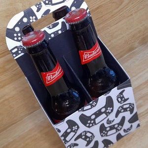 K&ouml;nnte beinhalten: Ein Sechs-Pack-Tr&auml;ger mit einem wei&szlig;en und schwarzen Muster von Videospielcontrollern. Der Tr&auml;ger ist gef&uuml;llt mit vier braunen Glasflaschen Budweiser-Bier.