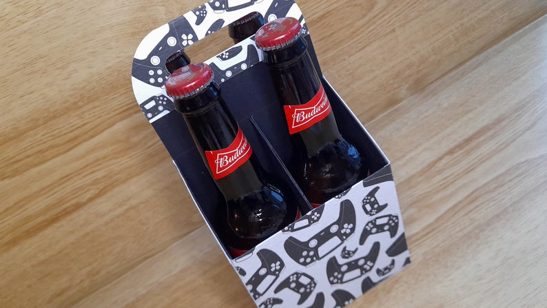 K&ouml;nnte beinhalten: Ein Sechs-Pack-Tr&auml;ger mit einem wei&szlig;en und schwarzen Muster von Videospielcontrollern. Der Tr&auml;ger ist gef&uuml;llt mit vier braunen Glasflaschen Budweiser-Bier.