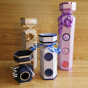 Puede incluir: Cuatro cajas de cartón decorativas con forma de cracker de Navidad. Las cajas están decoradas con cintas doradas, azules y moradas y tienen diferentes patrones. Las cajas están todas cerradas y listas para llenarse de regalos.