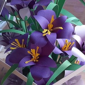 May include: A bouquet of purple and white paper flowers with yellow centers. The flowers are arranged in a clear glass vase. The flowers are made of paper and have a realistic appearance.
