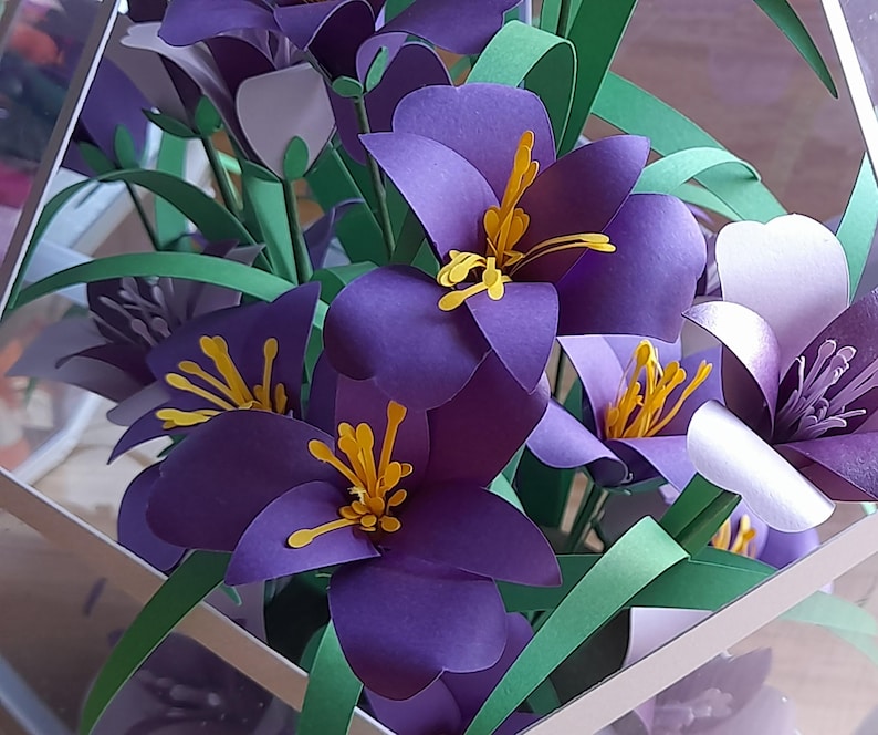 May include: A bouquet of purple and white paper flowers with yellow centers. The flowers are arranged in a clear glass vase. The flowers are made of paper and have a realistic appearance.