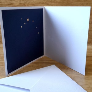May include: A white card with a navy blue inside featuring a gold star and smaller gold stars. The card is folded in half and is sitting on a wooden surface.