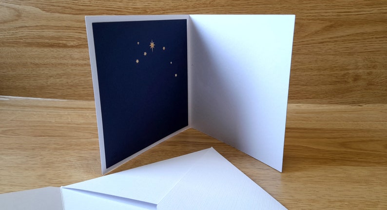 May include: A white card with a navy blue inside featuring a gold star and smaller gold stars. The card is folded in half and is sitting on a wooden surface.