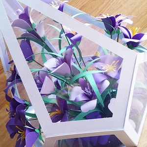 May include: A white paper geometric box with a clear plastic window, filled with a bouquet of purple and green paper flowers.