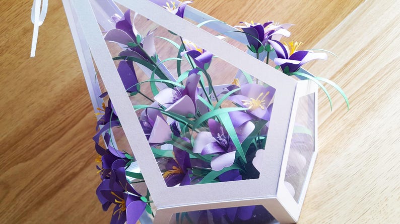 May include: A white paper geometric box with a clear plastic window, filled with a bouquet of purple and green paper flowers.