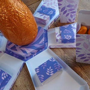 May include: A purple and white patterned explosion box with a chocolate egg in the center. The box has six compartments, each with a purple and white patterned flap. One compartment contains a small pile of orange candies.