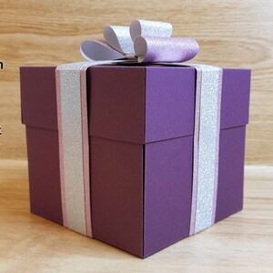 May include: A purple square-based explosion box with a silver and pink ribbon and bow. The box is decorated with silver glitter.  Sturdy square-based explosion box with faux ribbon and bow. Versatile gift box could be used to present lots of different things.