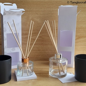 Può includere: Due bottiglie diffusori in vetro trasparente con bastoncini di legno, una con tappo in rame e l'altra con tappo in argento. Le bottiglie sono su basi bianche e sono accanto a due barattoli di candele neri. Le bottiglie sono davanti a due scatole bianche con una finestra per mostrare il liquido diffusore all'interno. Le scatole hanno un motivo floreale.